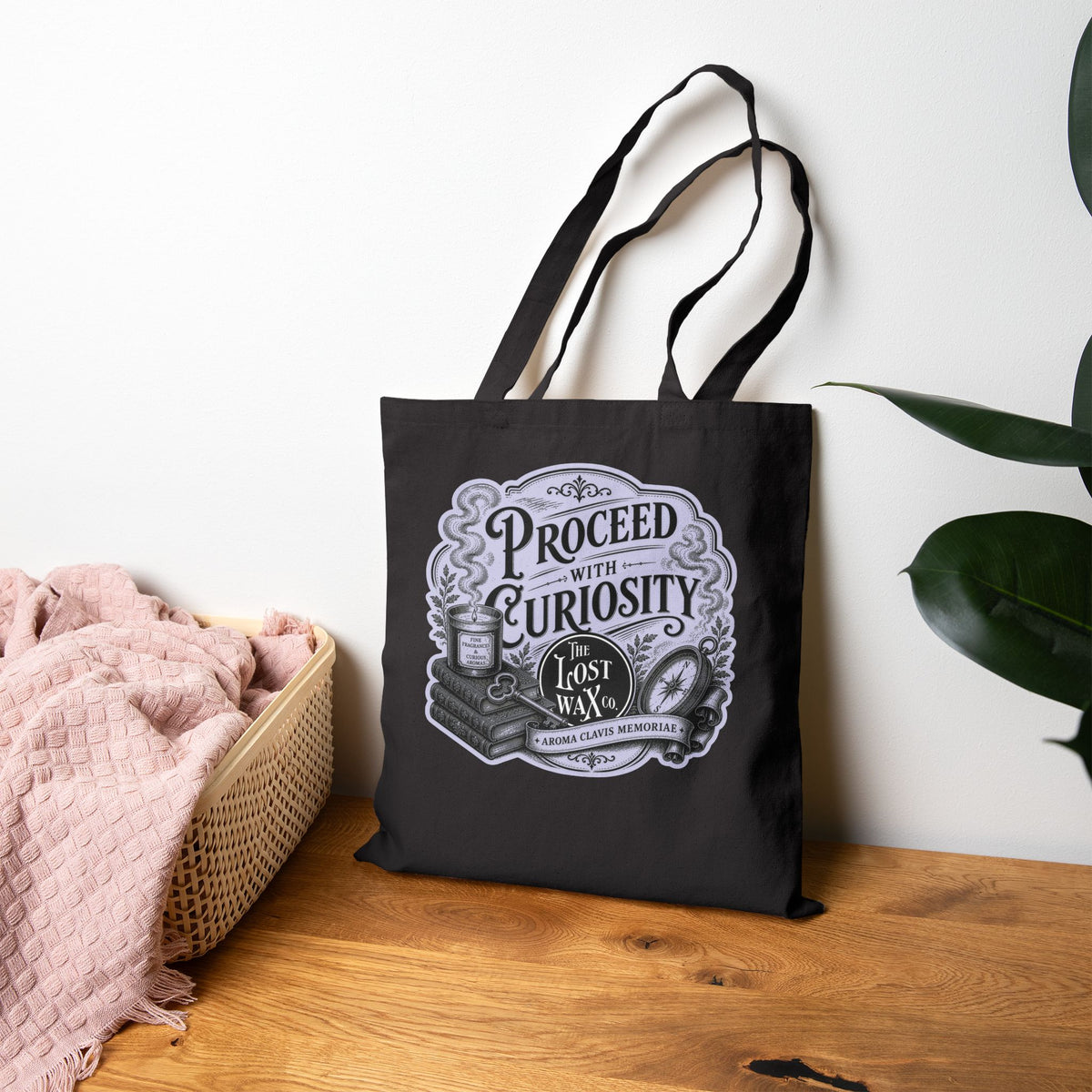 A black canvas tote bag with long handles stands on a wooden surface against a white wall. The bag has a large vintage-style lavender graphic print reading 'PROCEED WITH CURIOSTY' and 'THE LOST WAX CO.'