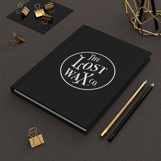 Black notebook with 'The Lost Wax Co.' logo on a dark surface with office supplies.
