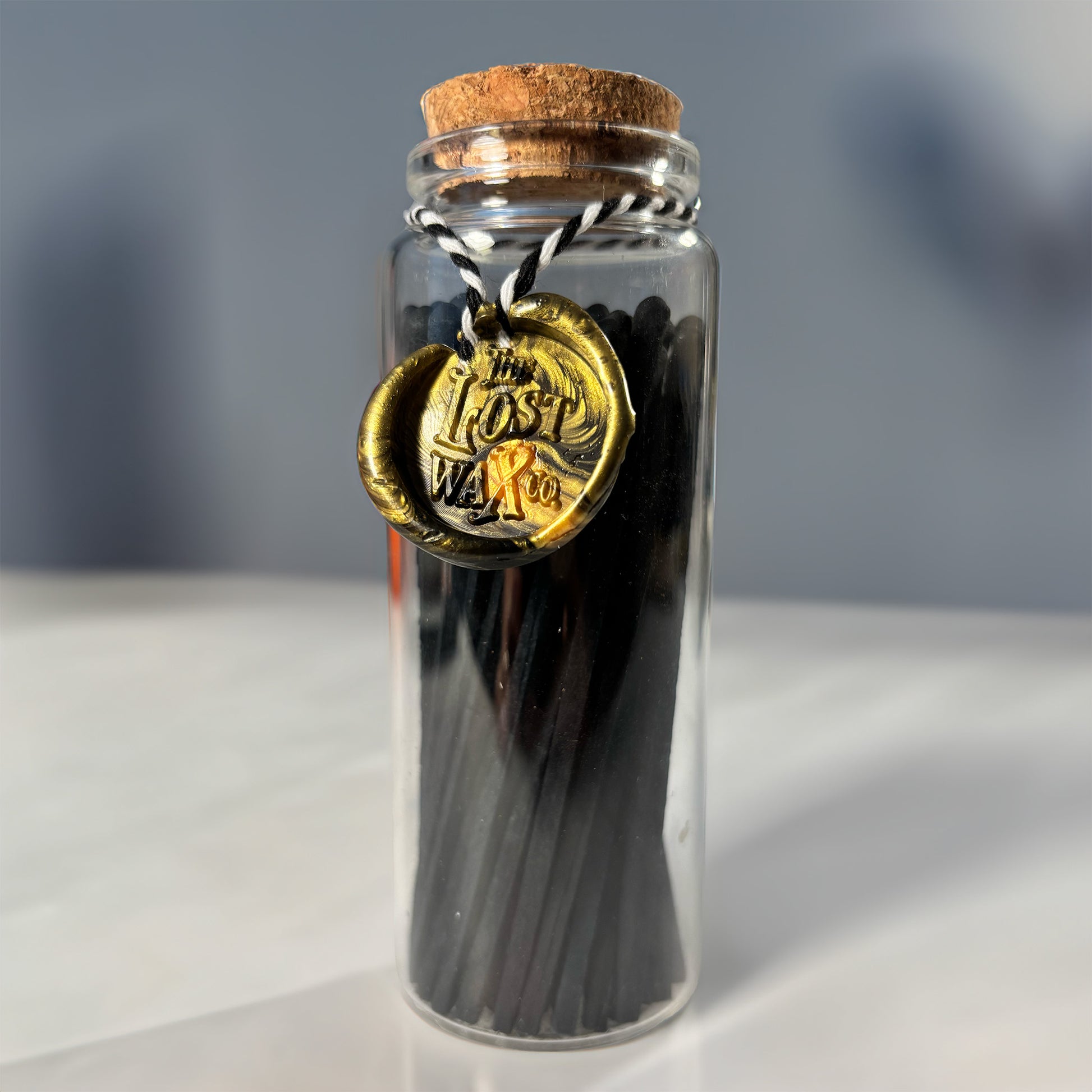 Glass jar with cork lid containing black safety matches with a gold seal labeled 'The Lost Weave Co.' on a neutral background.