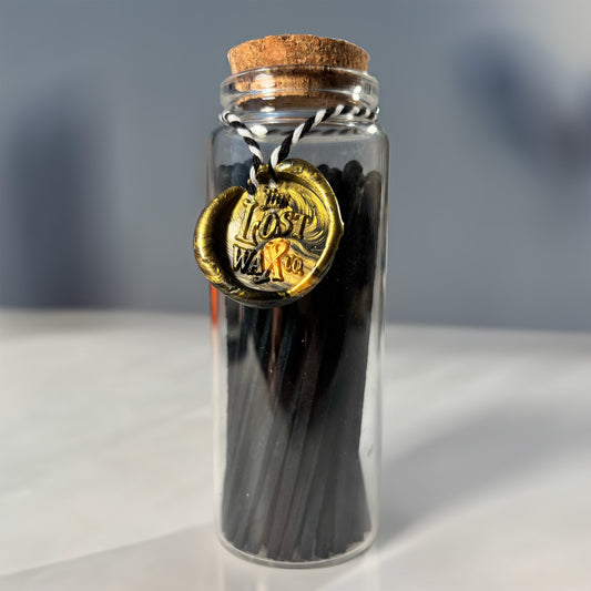 Glass jar with cork lid containing black safety matches with a gold seal labeled 'The Lost Weave Co.' on a neutral background.
