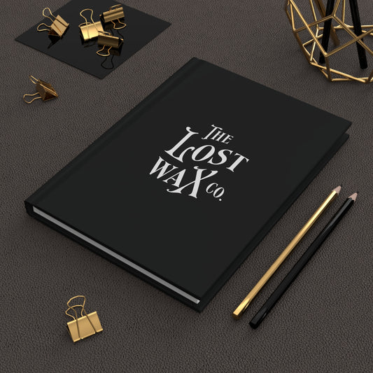 Black notebook with 'The Lost Wax Co.' on a dark surface with office supplies.