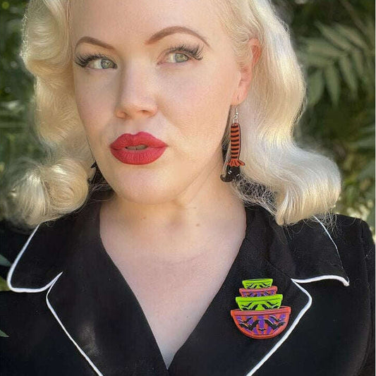 A close-up portrait of a woman with platinum blonde hair, wearing a black satin-like top with white piping and a colorful striped enamel lapel pin. She is looking to the side against a blurred green outdoor background.
