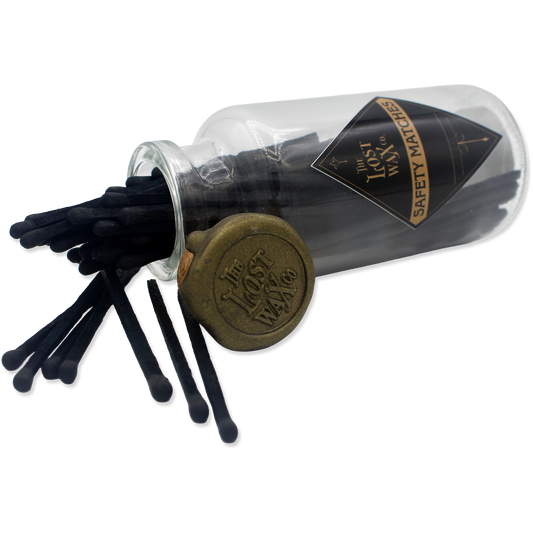 Glass jar with cork lid containing black safety matches labeled 'The Lost Wax Co.' on a white background.