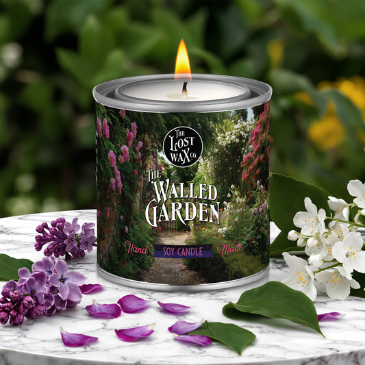 Candle in a decorative tin labeled 'The Walled Garden' with floral elements on a marble surface.