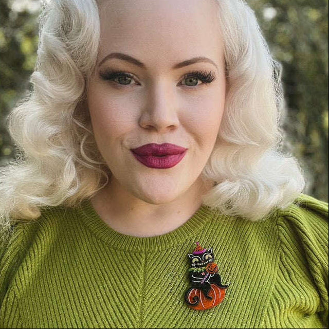 A woman with vintage-style platinum blonde hair and dark red lipstick wears a bright lime green ribbed sweater and a small Halloween-themed cat pin on the chest. The background is a soft-focus outdoor green setting.