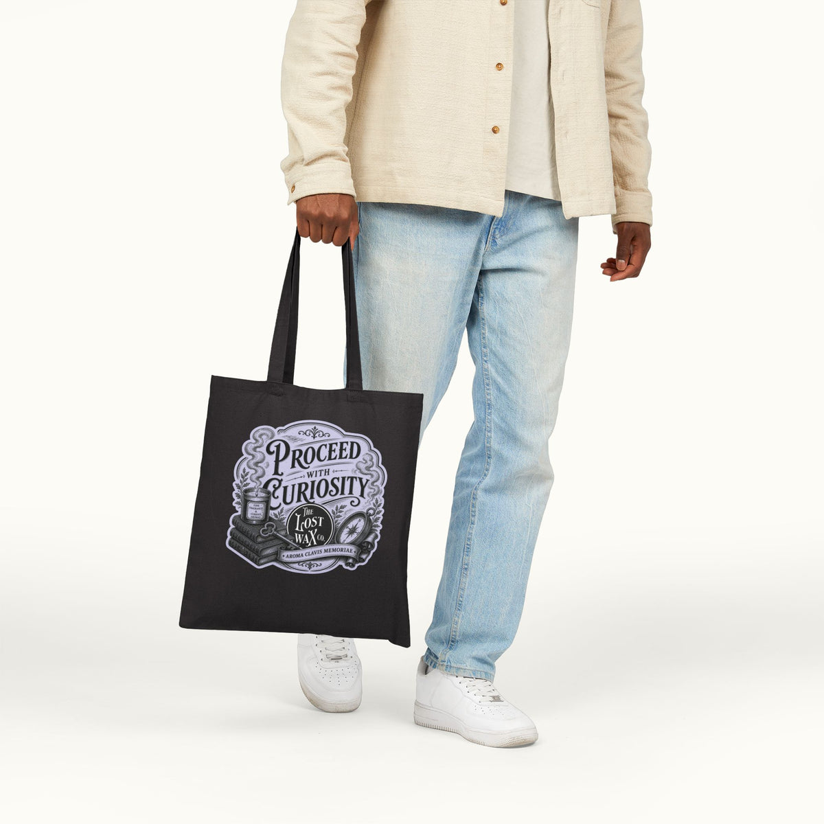 A person wearing a beige button-up shirt, light blue jeans, and white sneakers holds a black canvas tote bag with a vintage-style graphic that reads 'PROCEED WITH CURIOSITY."