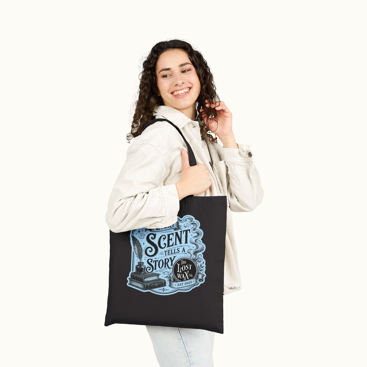 A woman wearing a light beige jacket and jeans holds a black canvas tote bag with a blue and black graphic that reads 'EVERY SCENT TELL A STORY' and 'The Lost Wax Co..' The photo is taken against a clean, off-white background.