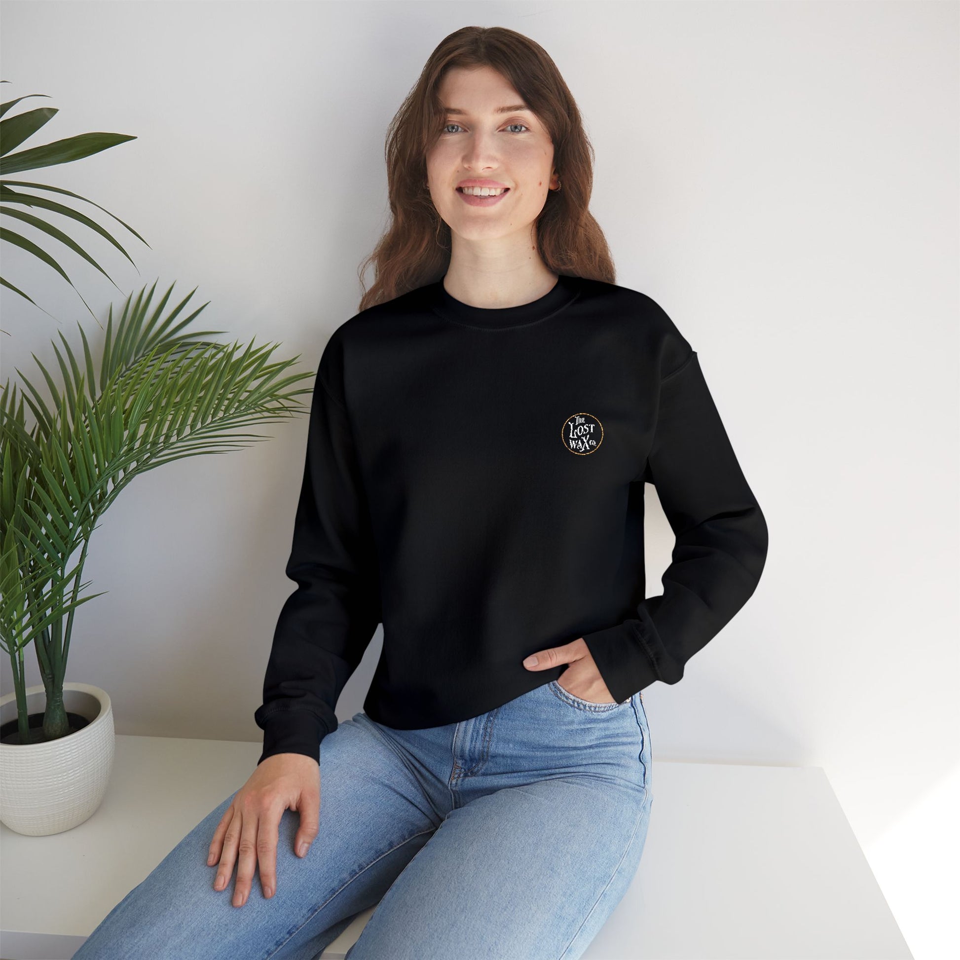 Woman wearing a black sweatshirt with a logo, sitting on a white surface with a plant in the background.