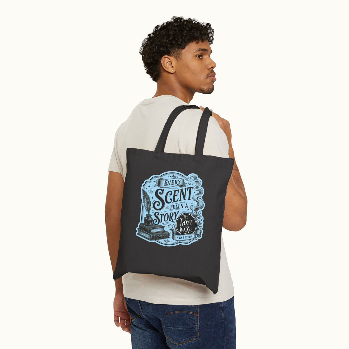 A man wearing a white t-shirt and blue jeans is shown from the back, carrying a black canvas tote bag over his shoulder. The bag features a light blue vintage-style graphic with the text 'EVERY SCENT TELLS A STORY' and illustrations of books and a quill pen.