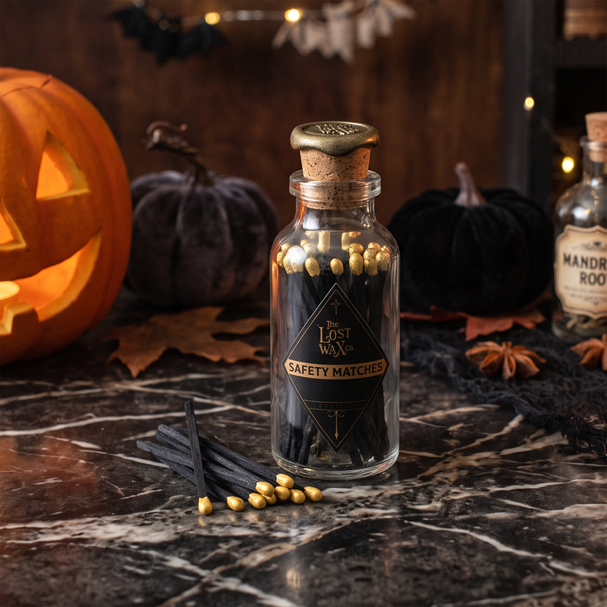 A glass bottle of 'The Lost Wax Co.' Black & Gold Safety Matches styled on a rustic table with Halloween props like a lit candle, a jack-o'-lantern, and old books.