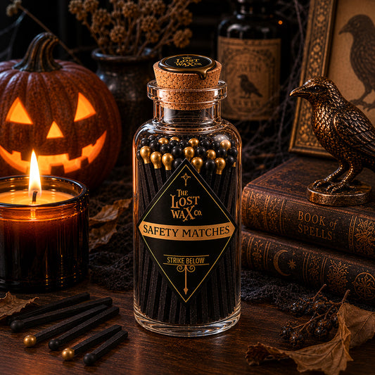 A glass bottle of 'The Lost Wax Co.' Black & Gold Safety Matches styled on a rustic table with Halloween props like a lit candle, a jack-o'-lantern, and old books.
