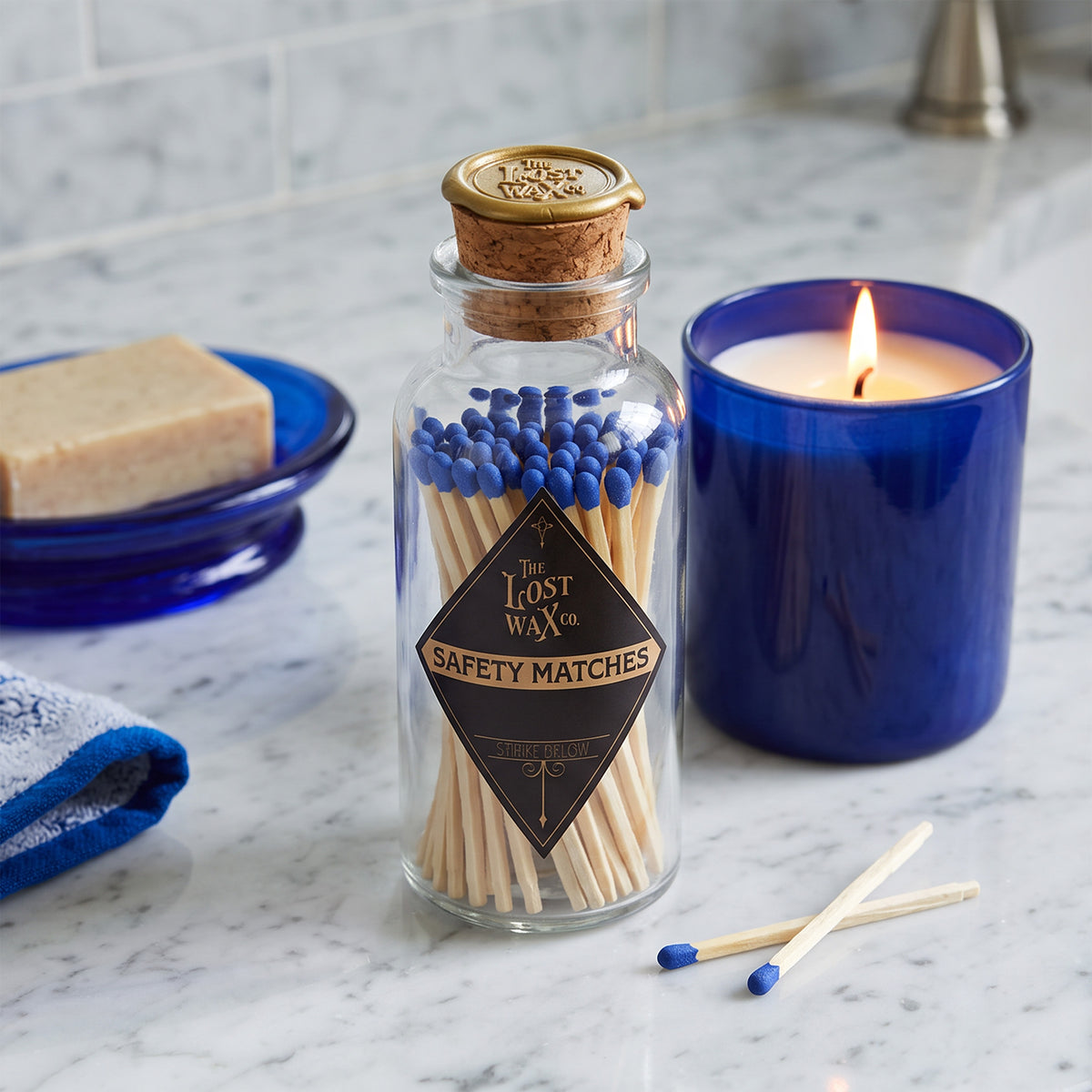 A glass jar with cork and wax seal containing 60 blue-tipped safety matches on a bathroom counter with blue jar candle, soap dish and blue towel.