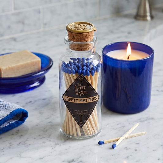 A glass jar with cork and wax seal containing 60 blue-tipped safety matches on a bathroom counter with blue jar candle, soap dish and blue towel.