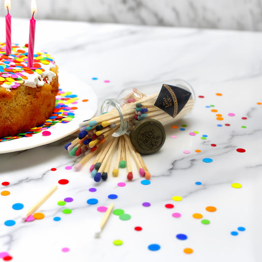 Glass bottle of colorful matches spilling out onto a marble counter with multi-color confetti, and a decorated cake with candles.