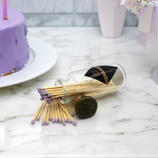 A clear glass bottle tipped on its side with wooden matches spilled out onto a white marble surface. The matches have light wood bodies and lavender-purple tips.