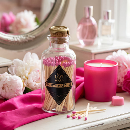 A clear glass bottle of safety matches with pink-tipped matches and a cork stopper, styled on a white surface with pink fabric, flowers, a pink candle, and perfume bottles in the background.