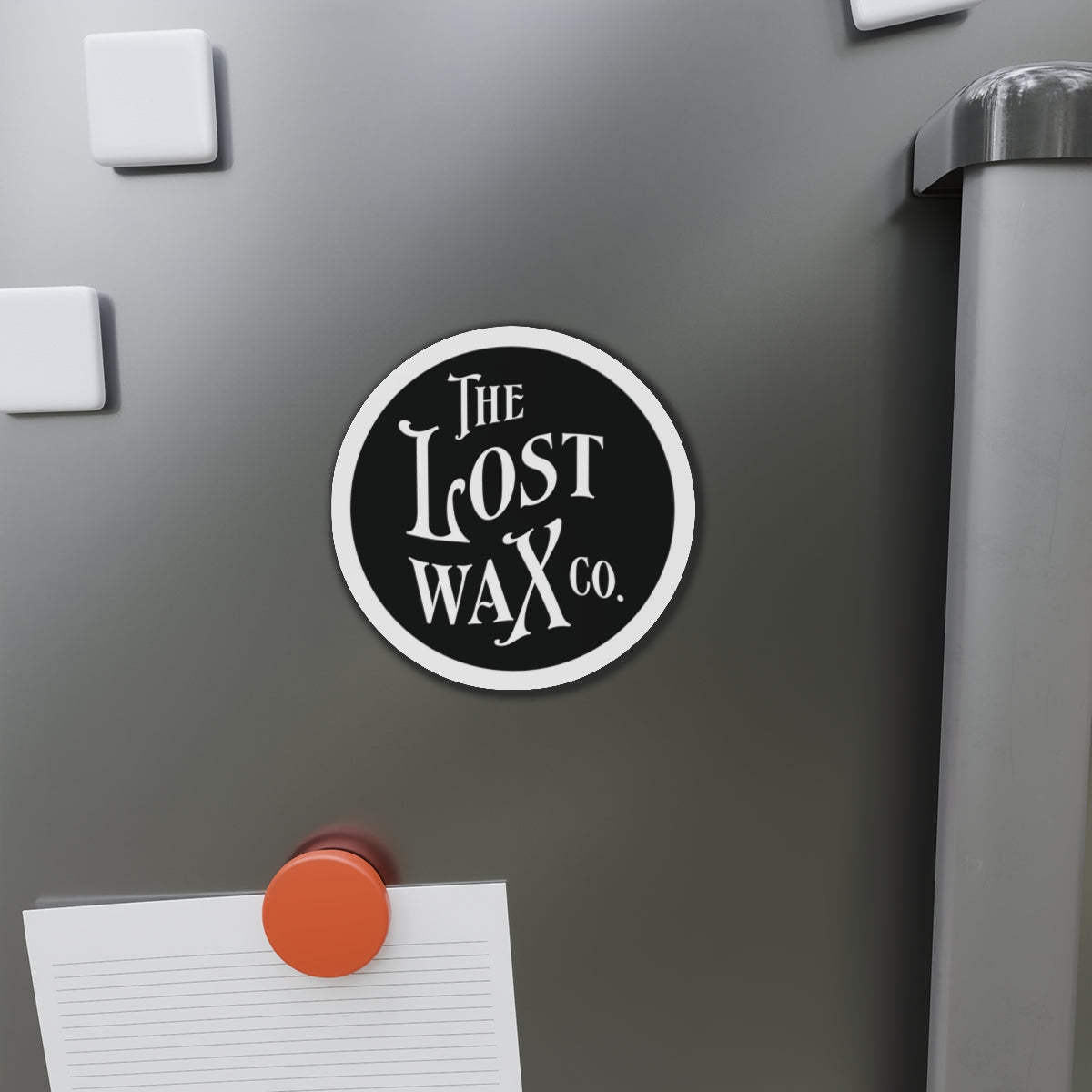 Round magnet with 'The Lost Wax Co.' on a refrigerator door.