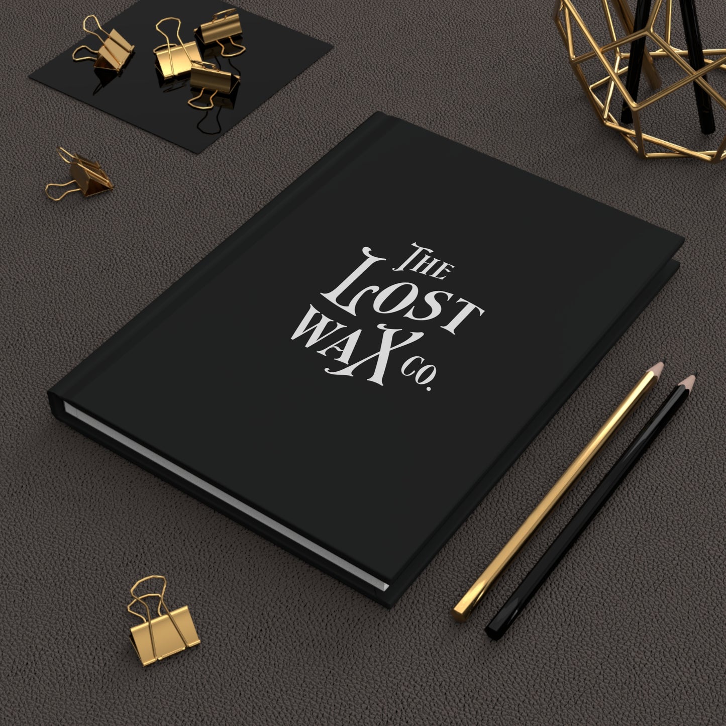 Black notebook with 'The Lost Wax Co.' on a dark surface with office supplies.