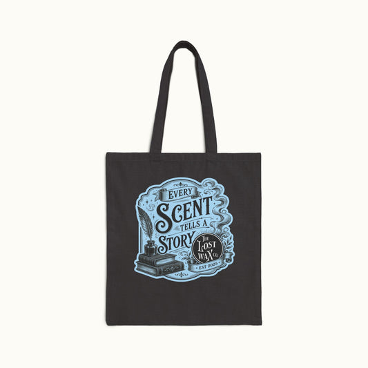 A black canvas tote bag with long handles, featuring a light blue and white vintage-style graphic on the front that reads 'EVERY SCENT TELLS A STORY' and 'The Lost Wax', shown against a plain off-white background.
