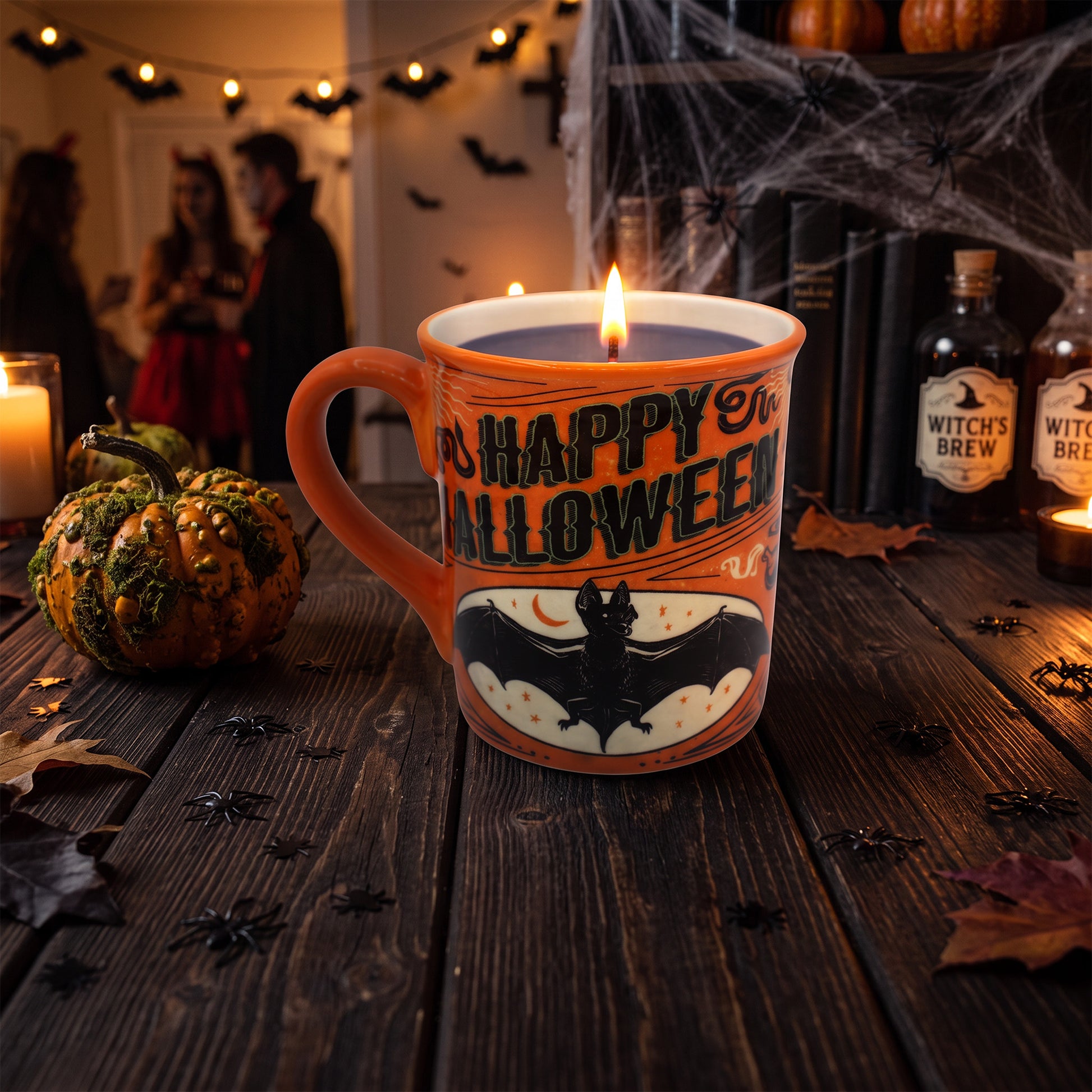 An orange ceramic candle mug with 'Happy Halloween' text and a bat graphic, lit on a dark wooden table with pumpkins, spiderwebs, and blurred people in the background.