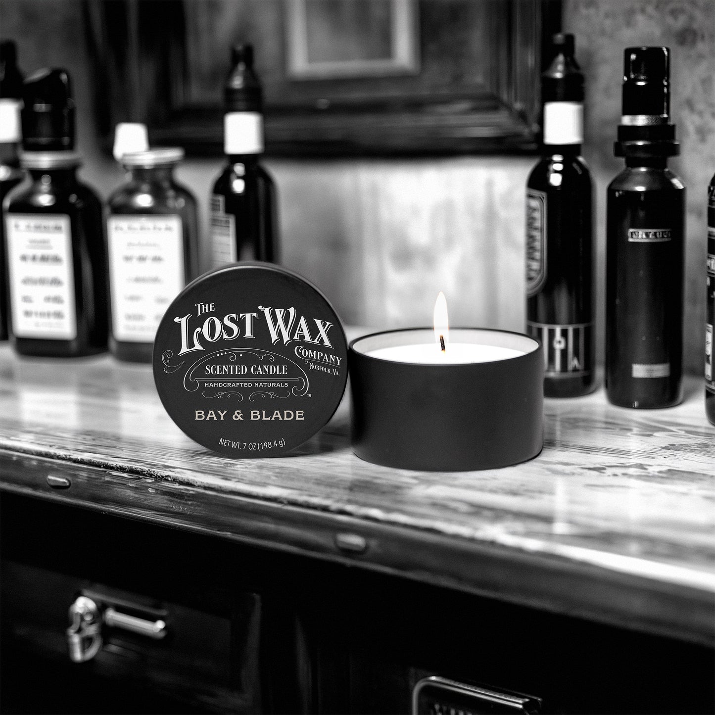 Candle and container labeled 'The Lost Wax Company' on a shelf with bottles in the background