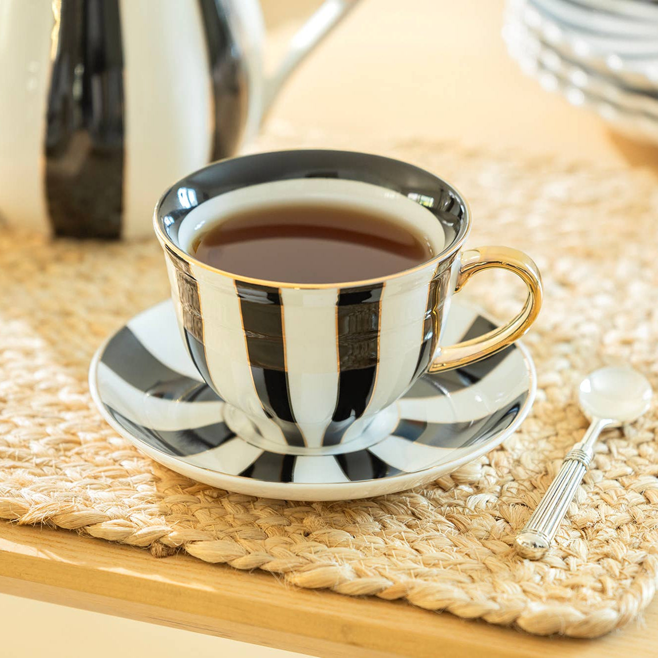 Black and white striped teacup with saucer on a woven mat, with a teapot and spoon in the background.
