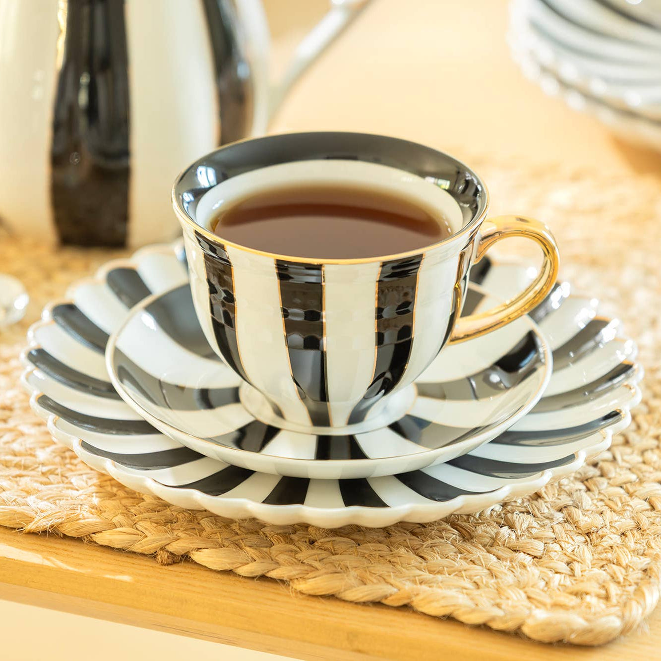 Black and white striped teacup and saucer on a woven placemat with a teapot in the background.