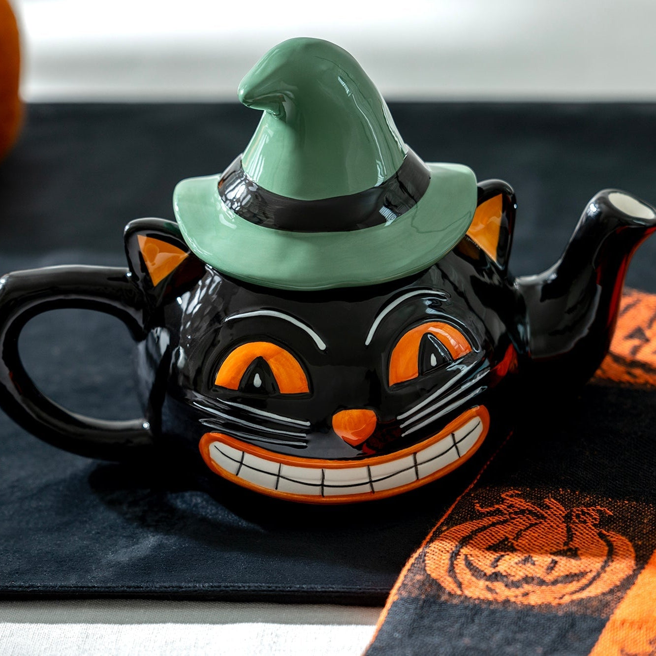 Halloween-themed black cat teapot with a witch hat on a tablecloth with pumpkins.