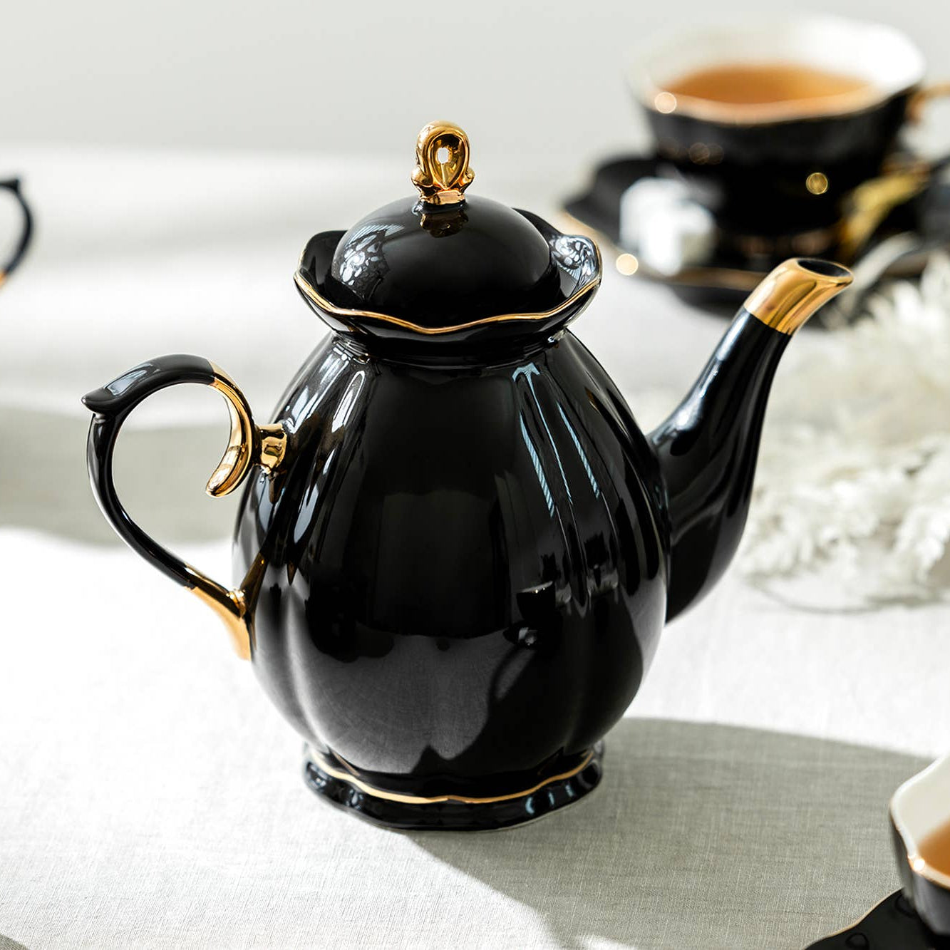 Black teapot with gold accents on a white surface with tea cups and saucers.