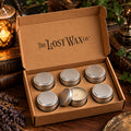 An open kraft cardboard gift box from The Lost Wax Co. containing six small silver metal tin candles, with five closed and one open showing the white wax.