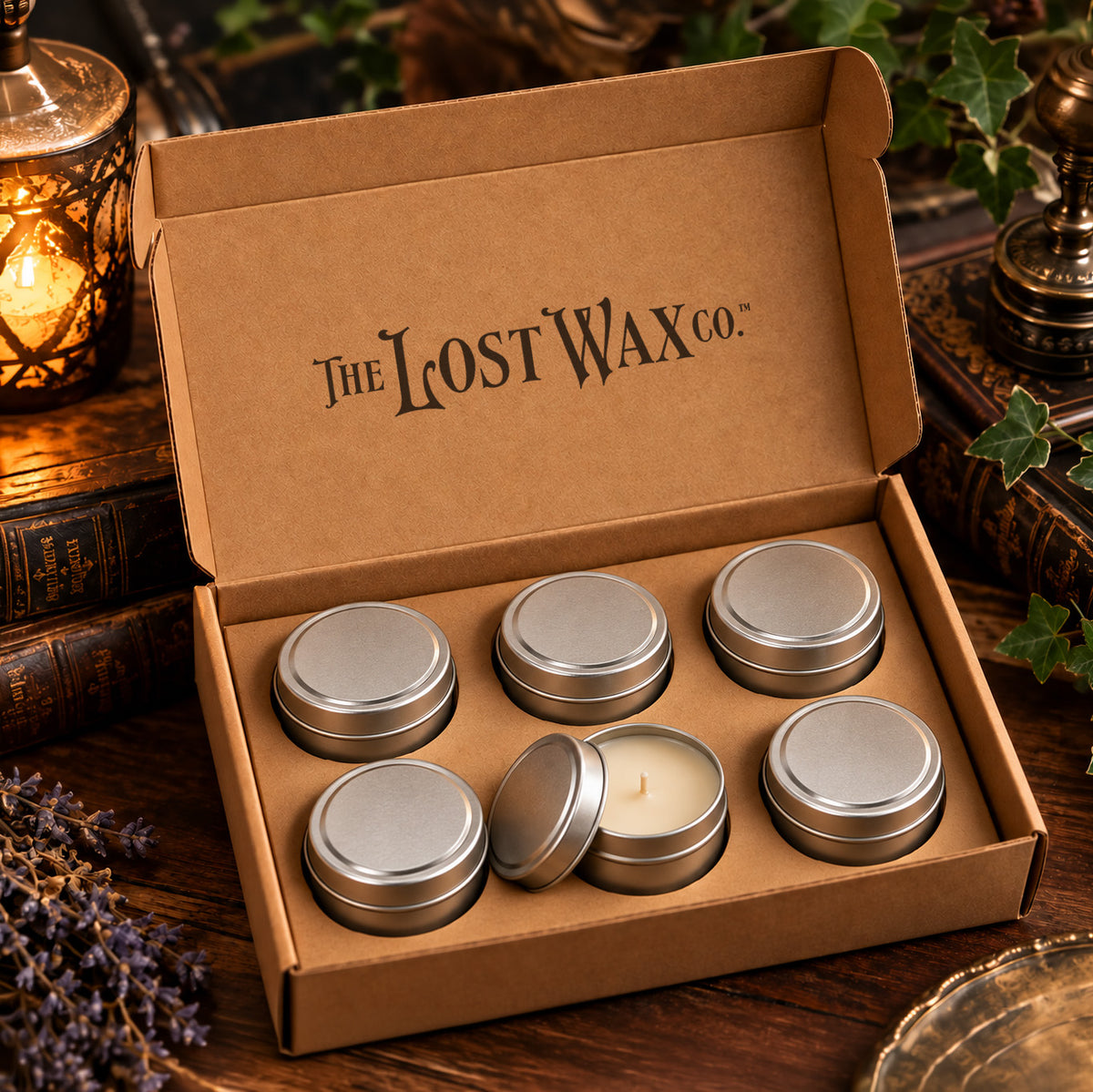 An open kraft cardboard gift box from The Lost Wax Co. containing six small silver metal tin candles, with five closed and one open showing the white wax.