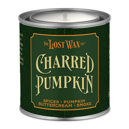 A single-wick candle in a metal tin canister with a dark green label reading 'The Lost Wax Co. Charred Pumpkin' and smaller scent notes 'Spices • Pumpkin • Buttercream • Smoke'.