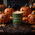 A lit candle in a green tin labeled 'The Lost Wax Charred Pumpkin' sits on a black lace tablecloth surrounded by carved pumpkins in a cozy indoor setting.