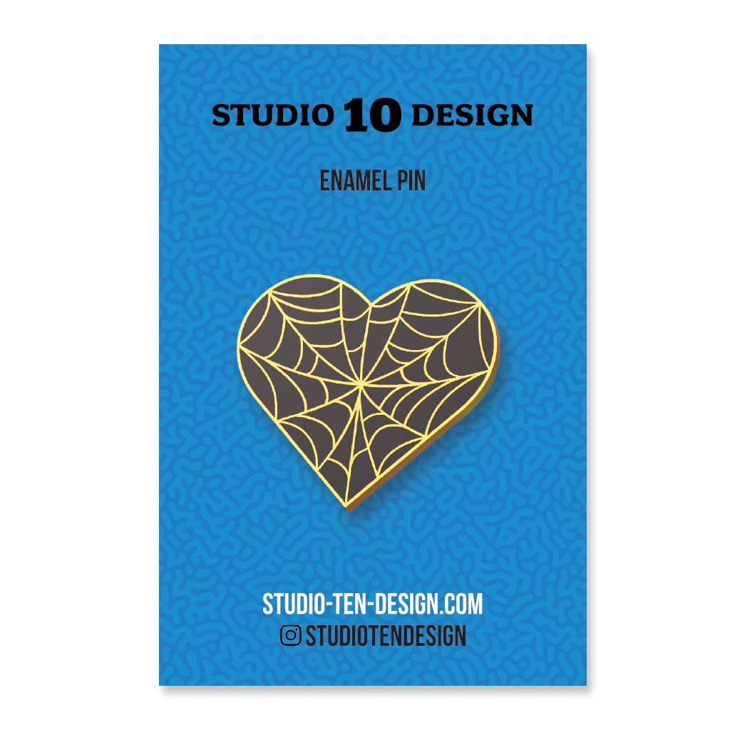 Heart-shaped enamel pin with spider web design on a blue card with 'Studio 10 Design' text.
