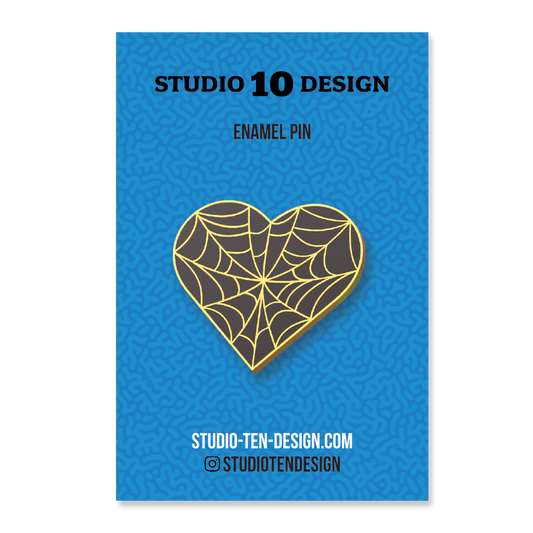 Heart-shaped enamel pin with spider web design on a blue card with 'Studio 10 Design' text.
