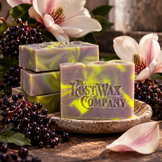 Bars of chartreuse and lavender soap stamped with "The Lost Wax Company" surrounded by elderberries and magnolia flowers.