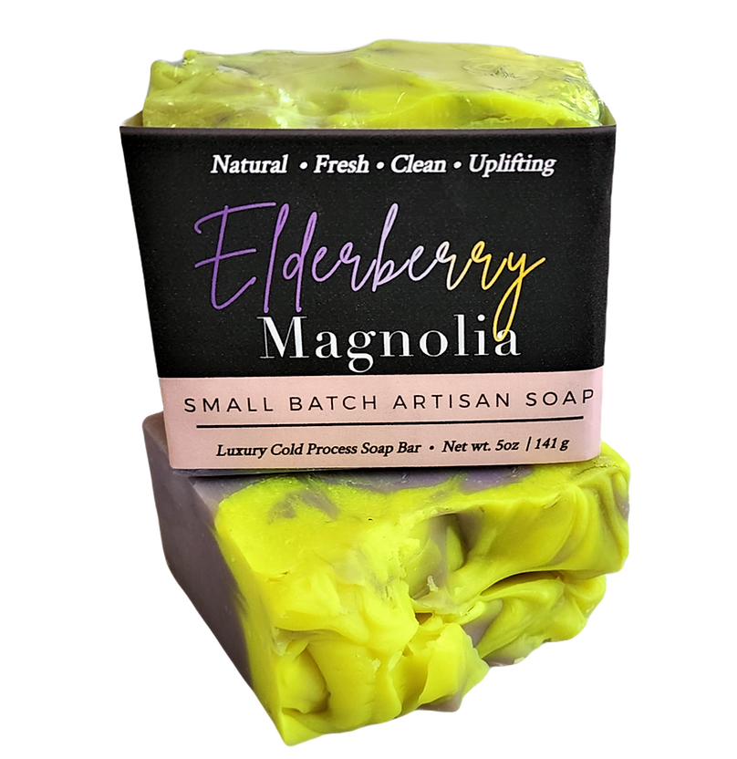 A bar of artisan soap labeled 'Elderberry Magnolia' with a black paper label, shown on a white background. The soap has a bright yellow-green top layer with darker marbling and a gray-purple side.