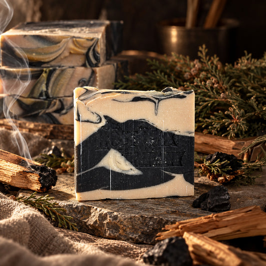 A bar of handmade black and white soap, surrounded by a rustic scene including a stick of burning palo santo wood, and cedar.