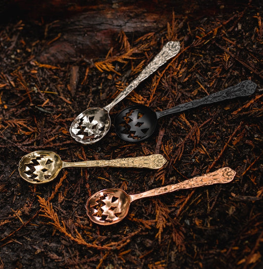 A group of four tea spoons with an ornate handle design, and jack-o-lantern cutouts in the bowl of the spoon. Silver, black, gold, and copper finishes.
