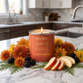 A lit LostWax 'Harvest Festival' scented candle sits on a marble kitchen counter surrounded by autumn flowers, pine branches, blackberries, and apple slices, with a blurred kitchen background.