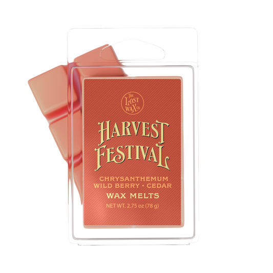 A package of Harvest Festival wax melts by The Lost Wax Co. The product is shown in its clear plastic clamshell packaging with a terracotta red label featuring gold lettering, with two reddish-orange wax melts visible behind it.