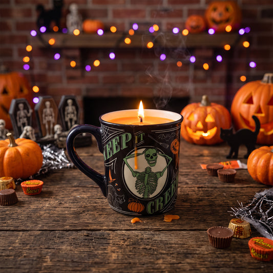 A Halloween-themed ceramic mug candle with a black handle and base, featuring a green skeleton graphic and the words 'KEEP IT CREEPY' on the front. The candle is lit and placed on a rustic wooden table surrounded by jack-o'-lanterns, candy corn, chocolate cups, and a black cat figurine, with a cozy fireplace and string lights in the background.