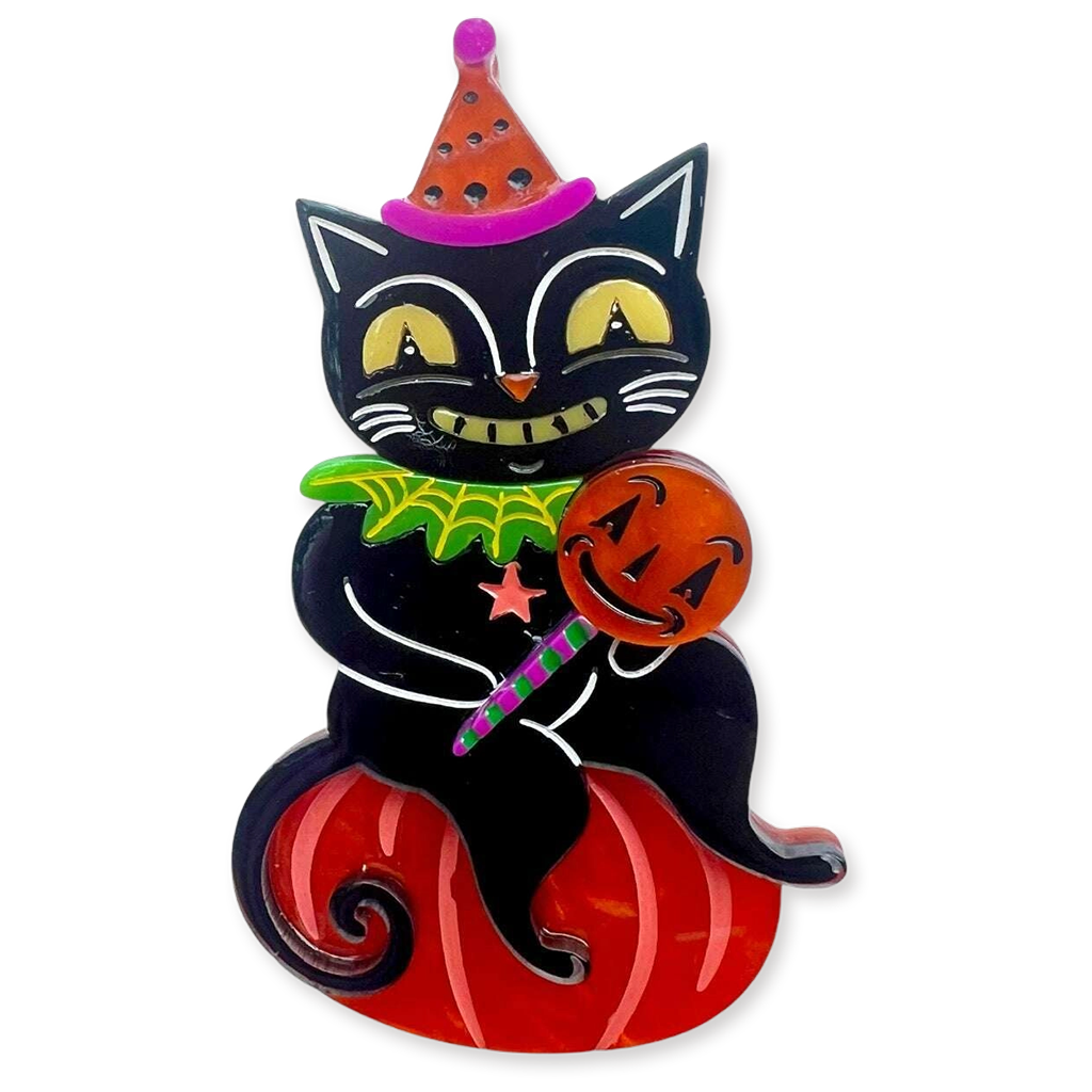 A Halloween-themed brooch featuring a black cat wearing an orange and pink pointed hat, holding a carved orange pumpkin, and sitting on a red pumpkin base. The cat has yellow eyes, a green collar with a spider detail, and holds a purple-striped candy cane.