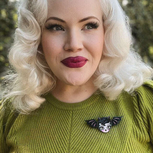 A close-up portrait of a woman with platinum blonde wavy hair wearing a textured olive green sweater and a small black-and-white bat brooch on the chest, set against a blurred outdoor background.