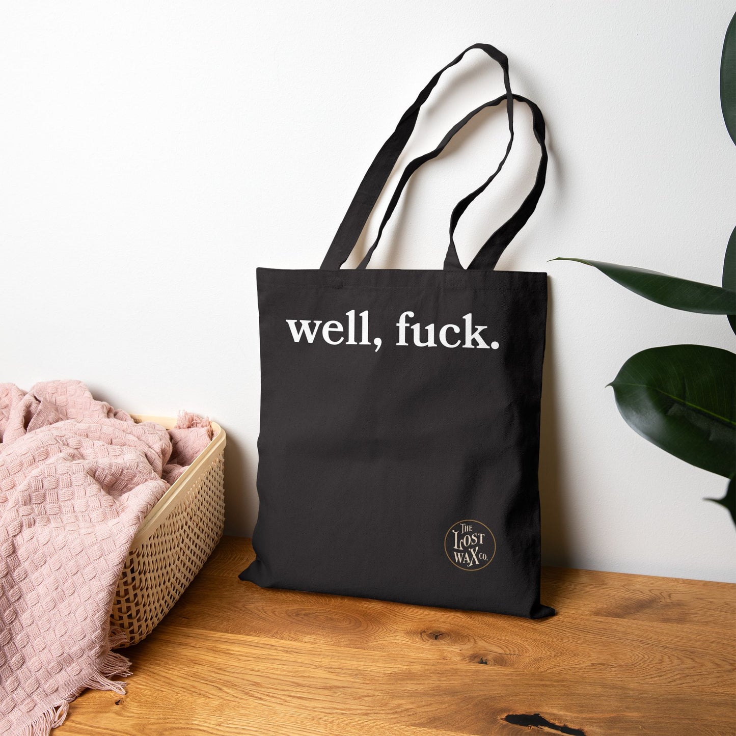 Black tote bag with 'well, fuck.' text on a wooden surface with a plant in the background