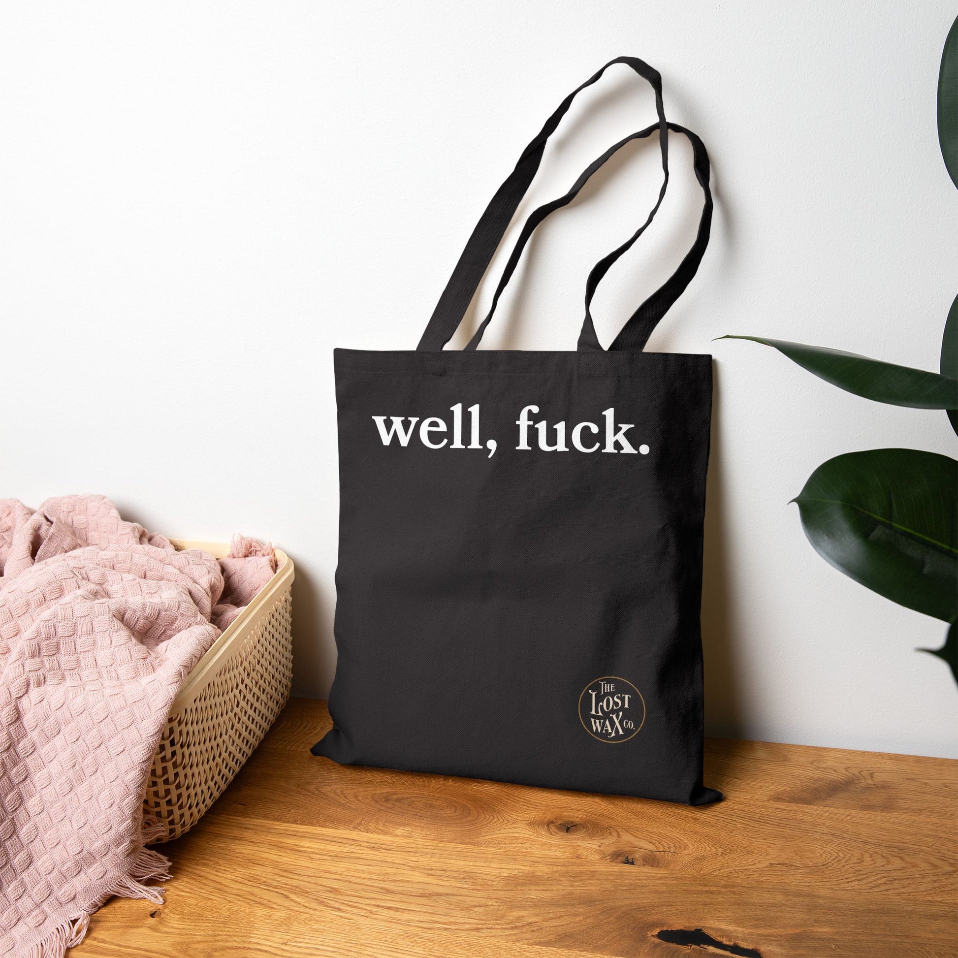 Black tote bag with 'well, fuck.' text on a wooden surface with a plant in the background