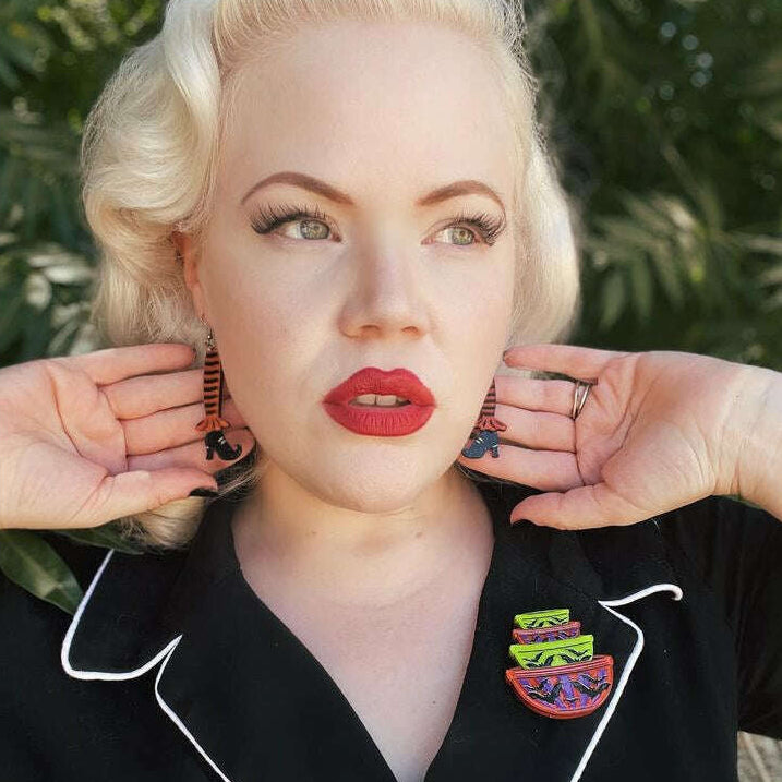 A close-up portrait of a woman with blonde hair wearing a black collared blouse with white piping and a colorful enamel pin, posed outdoors with her hands near her ears.