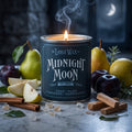 A lit candle in a dark teal tin labeled 'The Lost Wax Co. Midnight Moon' sits on a marble surface surrounded by pears, plums, cinnamon sticks, sandalwood, and sugar crystals in a moody nighttime setting.