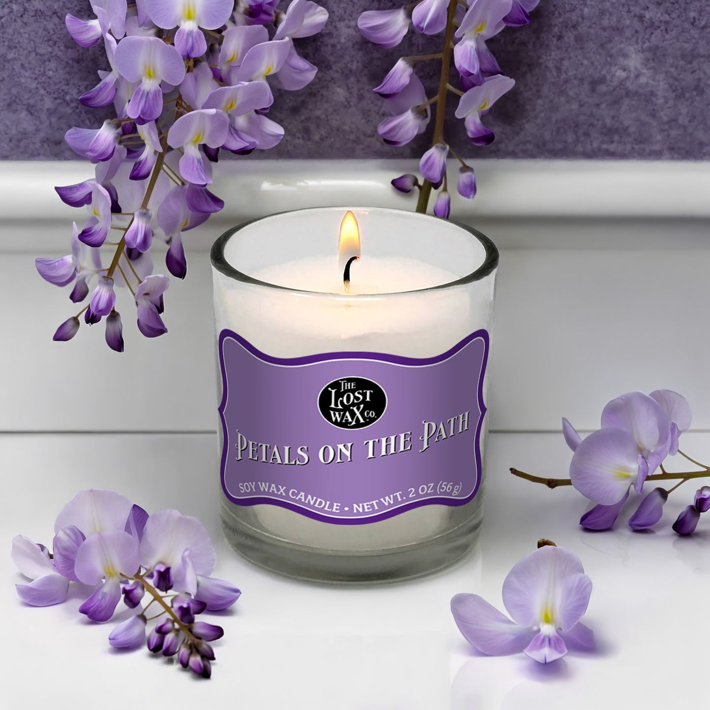 Candle with a purple label surrounded by purple flowers on a white surface