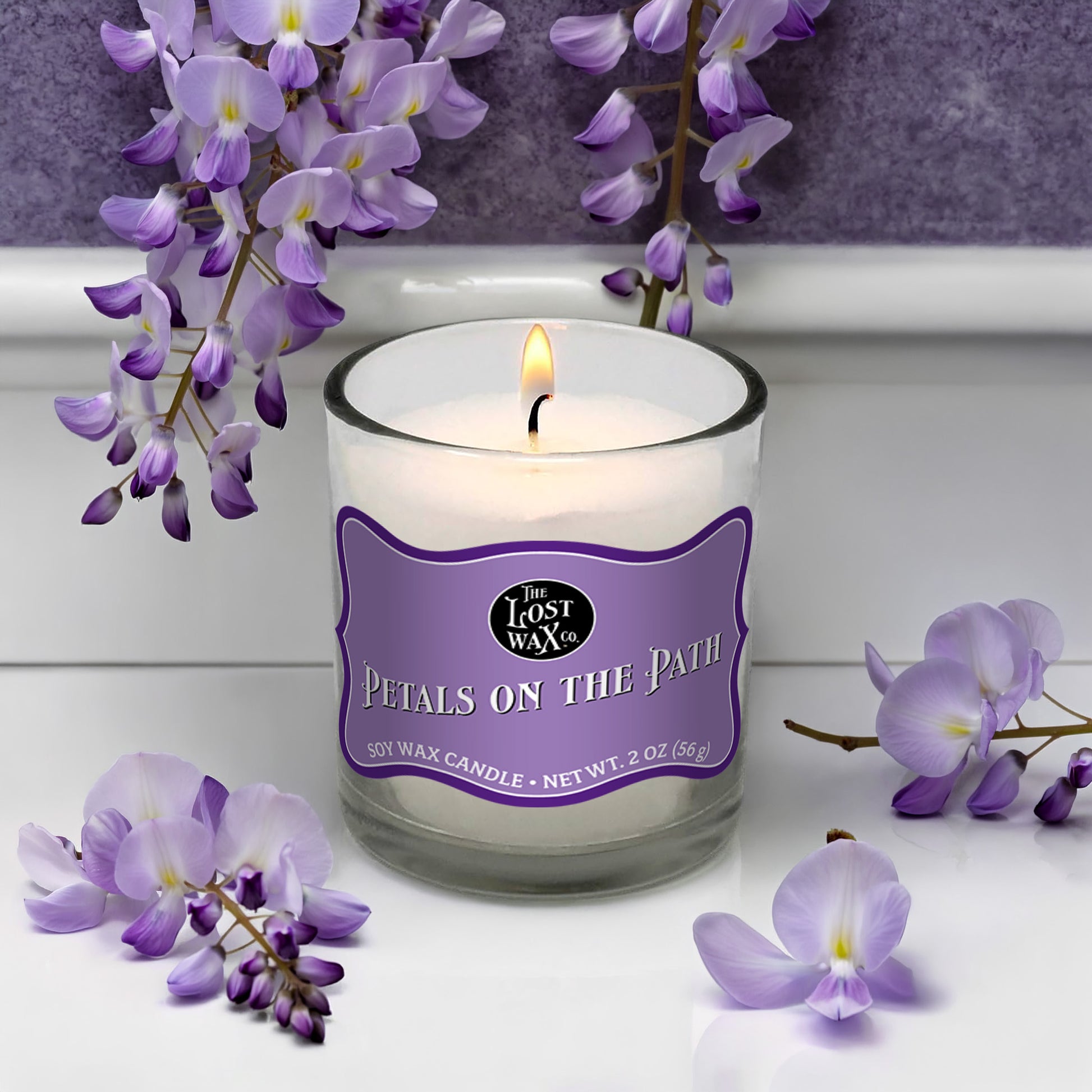 Candle with a purple label surrounded by purple flowers on a white surface